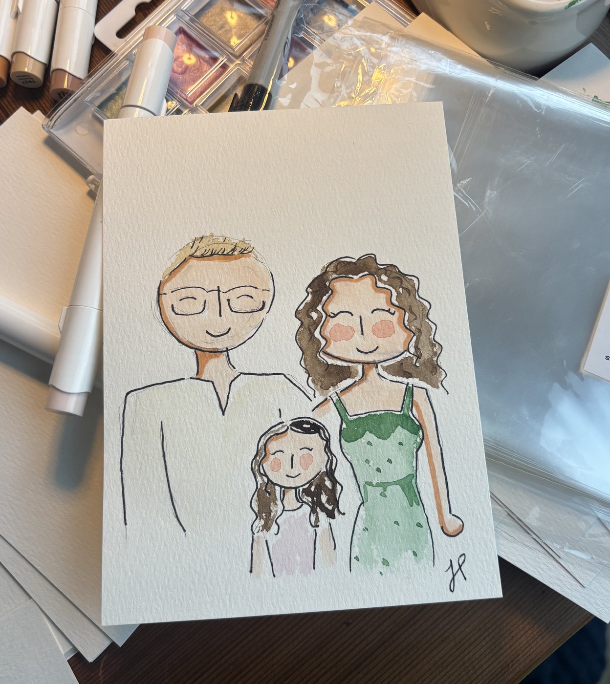 Watercolor portrait of a family of three, a dad in glasses, a mom in a green dress and their daughter, painted on watercolor paper at the studio desk