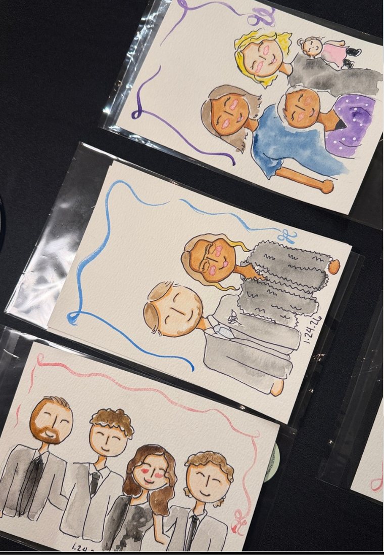 Three finished live guest portraits sleeved and laid out on a table: a couple beside their tiered wedding cake, a family group with a baby, and four groomsmen in suits, each painted in watercolor with a looping ribbon border and dated