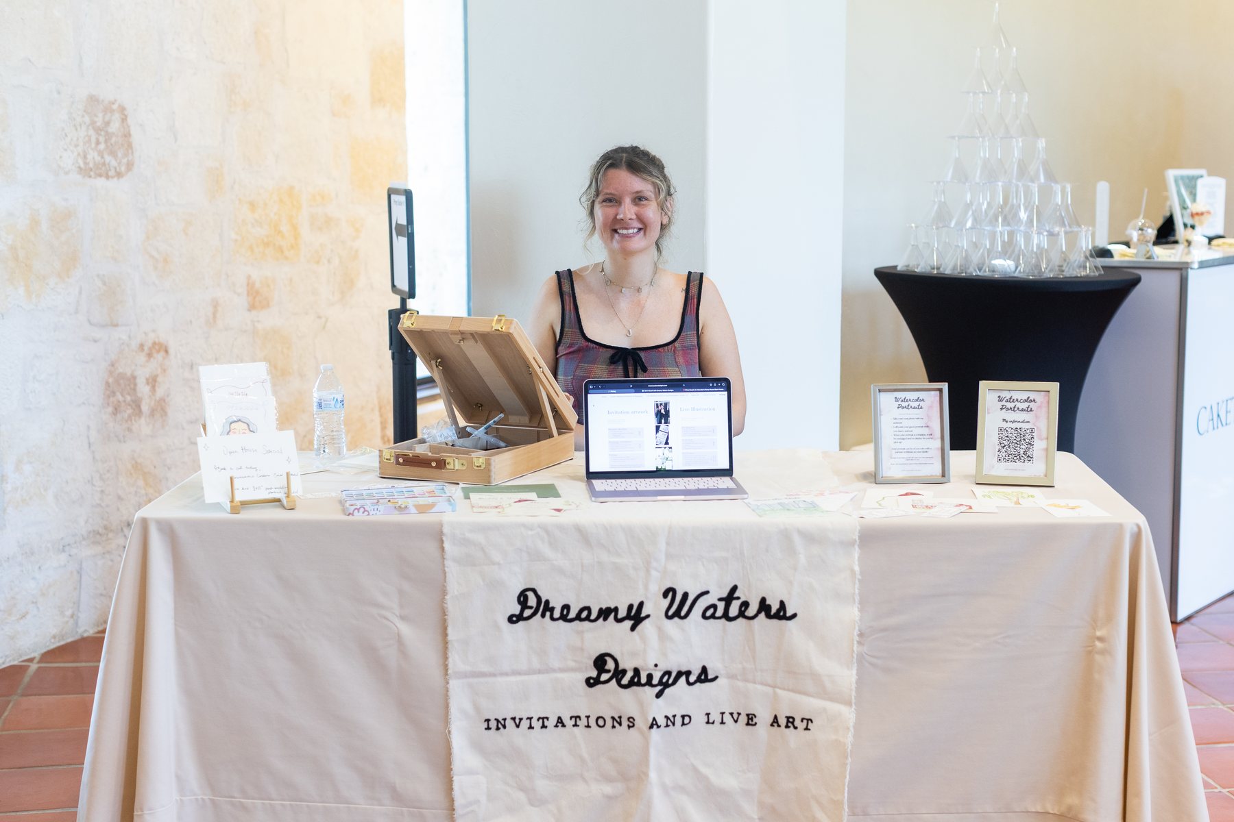 Jamielee behind her Dreamy Waters Designs table at a wedding venue, with her paint case open, framed watercolor portrait samples on display and an embroidered tablecloth reading Dreamy Waters Designs, Invitations and Live Art