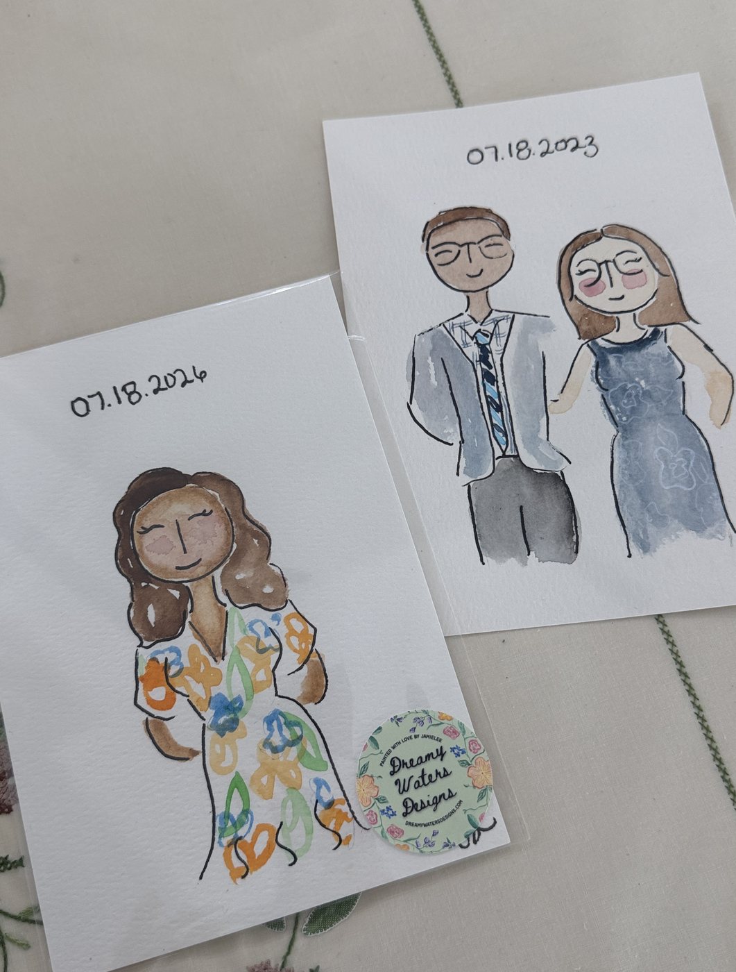 Two live guest portraits side by side: a woman in a floral dress dated 07.18.2026 with a Dreamy Waters Designs sticker, and a couple dated 07.18.2023
