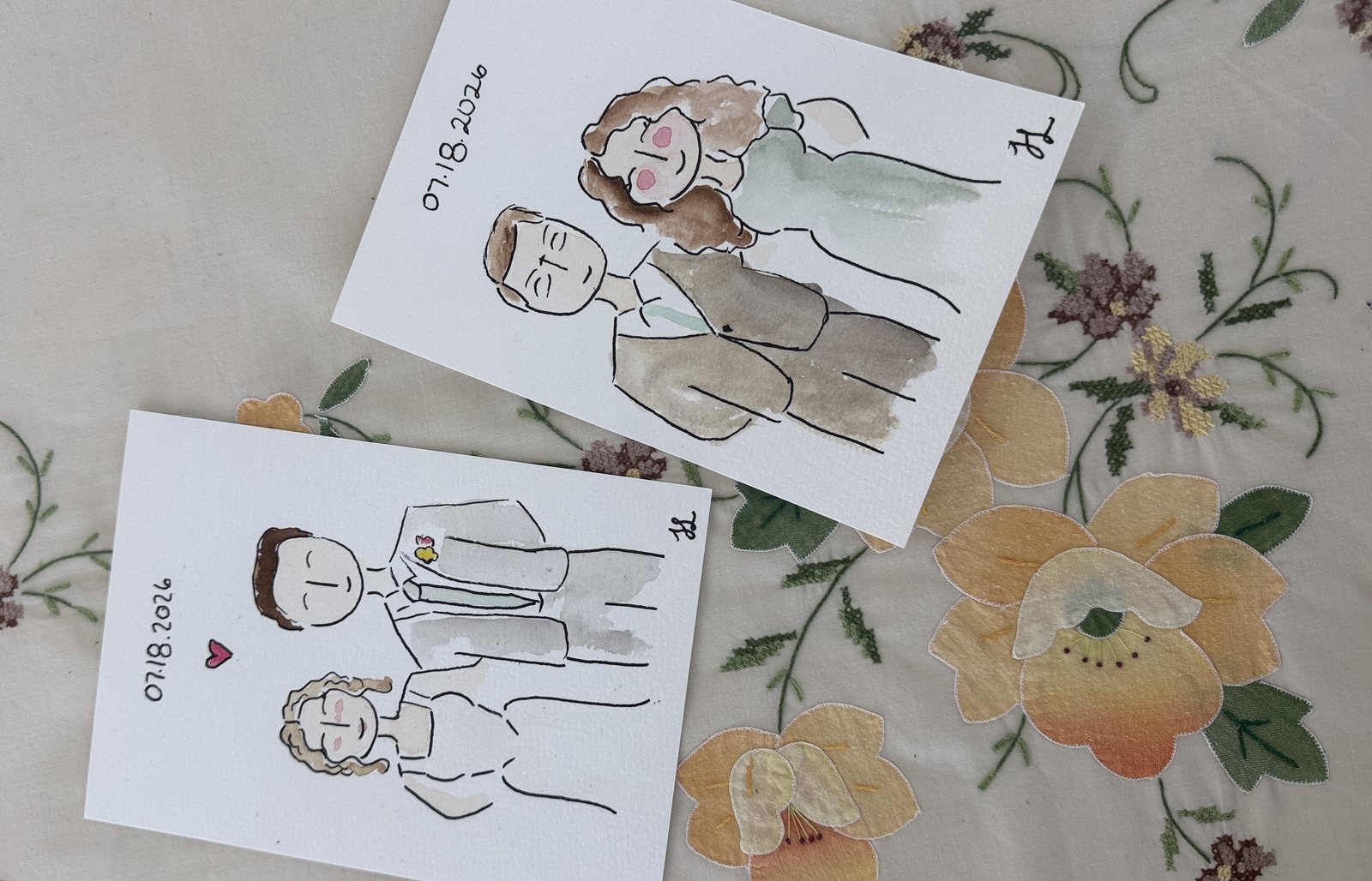 Two finished live guest portraits on a floral tablecloth: a couple dated 07.18.2026 and a groom in a light suit with a boutonniere